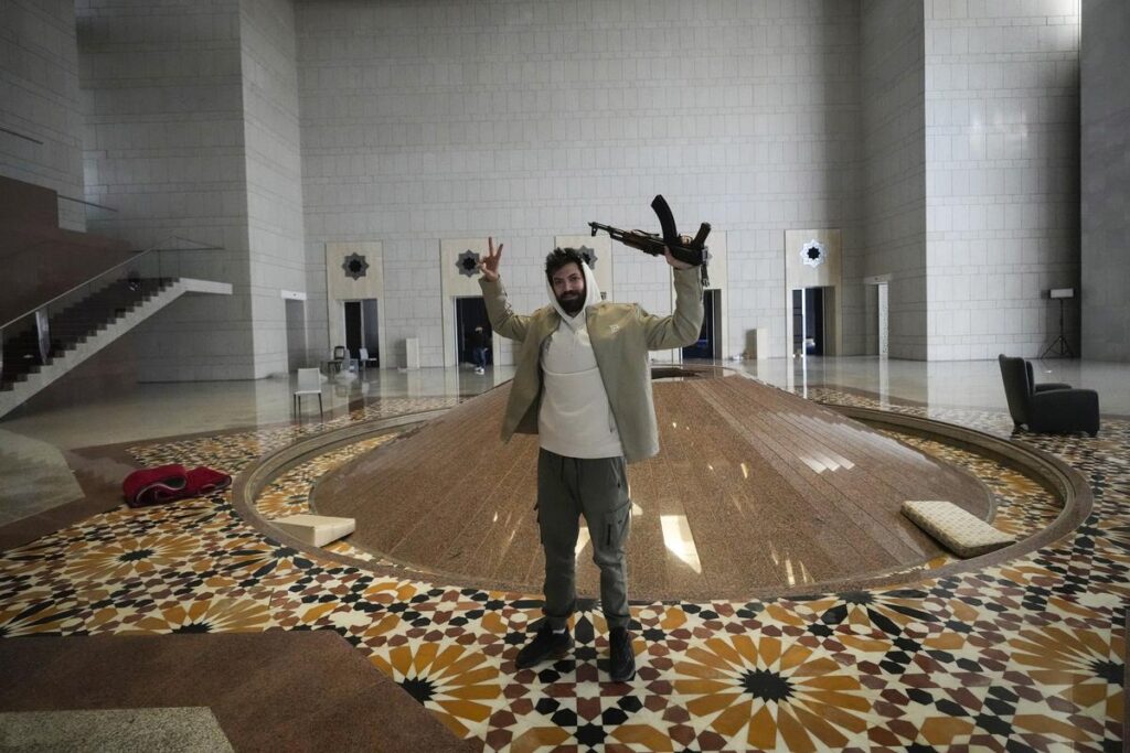 A man holds an AK-47 and flashes the victory sign as he poses for a photo in a hall of Syrian President Bashar Assad's presidential palace in Damascus, Syria, on December 8, 2024. | Photo Credit: AP